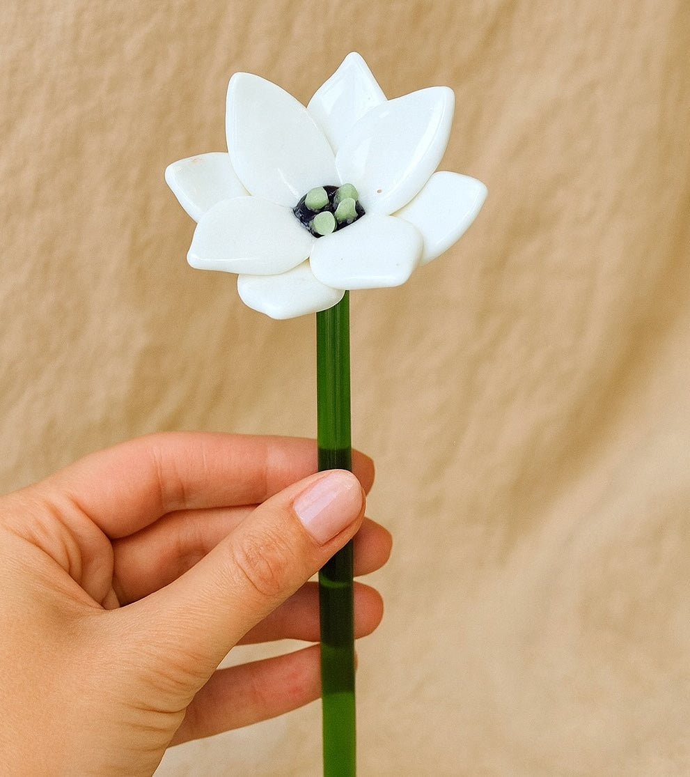 Hand holding a white ceramic flower with a green stem against a beige background