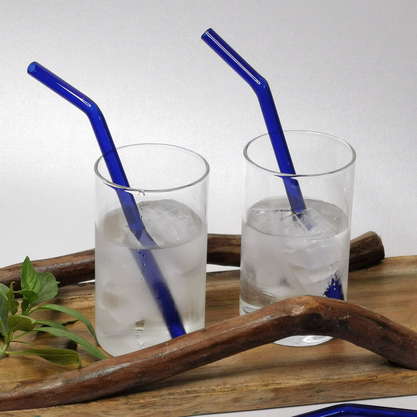 Two clear glasses filled with ice cubes and a blue glass straw in each, presented on a wooden surface with a cleaning brush lying beside them.
