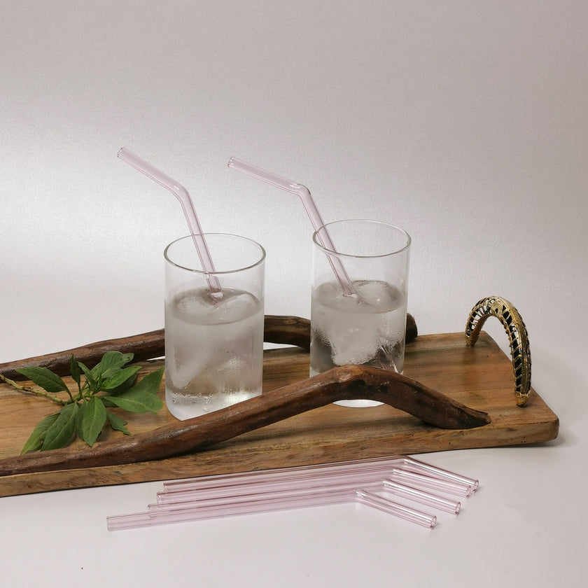 A set of pink glass straws displayed in a transparent glass with ice, presented on a wooden tray with a cleaning brush in the background.