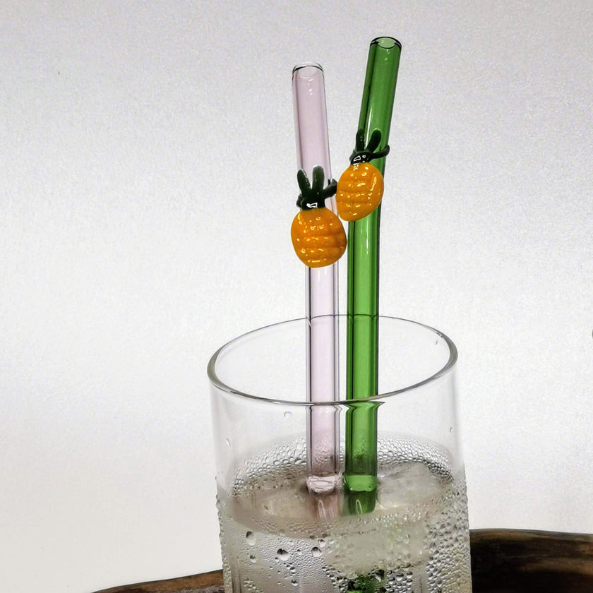 Two glass straws with pineapple decorations in a glass of water on a white background