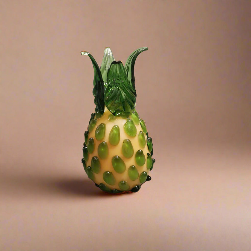 Handblown glass pineapple decoration with a green top, beige body and black accents on a grey background.