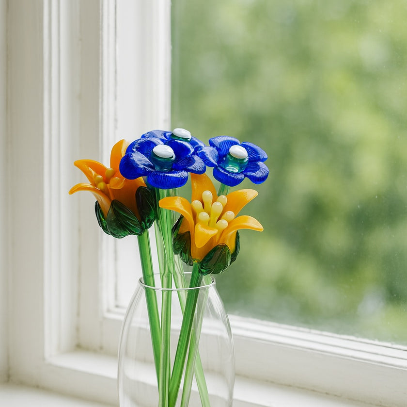 Colorful glass flowers in a clear vase by a window with greenery outside