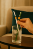 Hand holding a glass of water with a straw and decorative elements on a table.