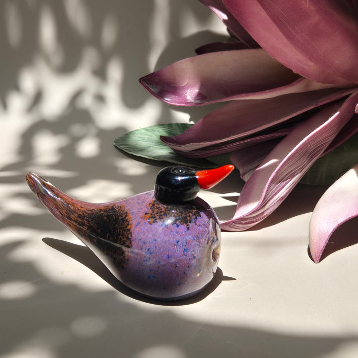 A handcrafted glass bird figurine with a red beak and swirls of violet, amber, and black colors, placed on a surface with a shadow of a plant leaf.